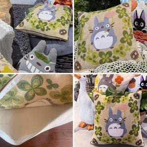 Puede incluir: Cojines decorativos cuadrados con dise&ntilde;os bordados. Un coj&iacute;n presenta un personaje Totoro gris rodeado de tr&eacute;boles verdes. Otro coj&iacute;n tiene un dise&ntilde;o de Totoro con una flor rosa. Los cojines est&aacute;n sobre un fondo beige.