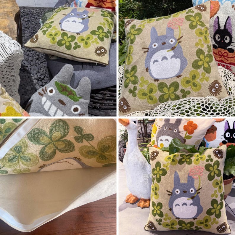 Puede incluir: Cojines decorativos cuadrados con dise&ntilde;os bordados. Un coj&iacute;n presenta un personaje Totoro gris rodeado de tr&eacute;boles verdes. Otro coj&iacute;n tiene un dise&ntilde;o de Totoro con una flor rosa. Los cojines est&aacute;n sobre un fondo beige.