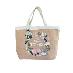 Puede incluir: Bolso tote beige con asas y ribetes blancos. El bolso presenta una corona floral bordada con el texto "My Neighbor Totoro" y un personaje de Totoro. Un peque&ntilde;o dije de Totoro cuelga del asa.