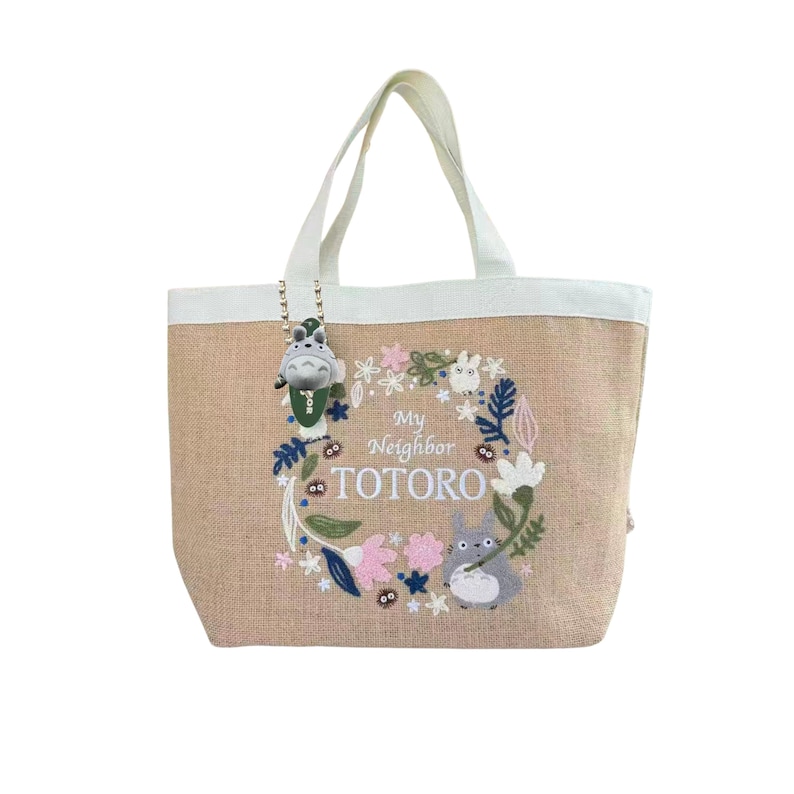 Puede incluir: Bolso tote beige con asas y ribetes blancos. El bolso presenta una corona floral bordada con el texto "My Neighbor Totoro" y un personaje de Totoro. Un peque&ntilde;o dije de Totoro cuelga del asa.