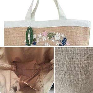 Puede incluir: Un bolso tote beige con asas y ribetes blancos. El bolso presenta un dise&ntilde;o floral bordado con un personaje de dibujos animados y el texto "My Neighbor Totoro". El interior es de color beige claro.