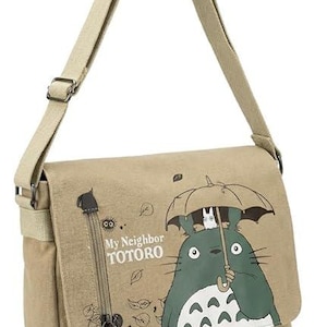 May include: Beige messenger bag featuring a green and white cartoon character holding an umbrella. The bag has a long, adjustable strap and a flap closure. The text "My Neighbor Totoro" is printed on the front.