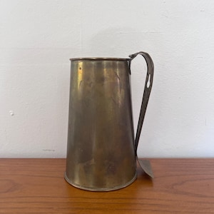 May include: An antique brass beer mug with a handle and a flared body. The mug has a tarnished, aged appearance. It is positioned on a wooden surface against a white wall, suggesting it is vintage.