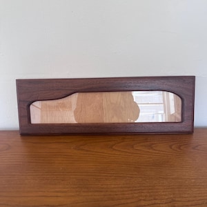 Può includere: Una porta di un mobile rettangolare in legno marrone scuro con un inserto in vetro trasparente. L'inserto in vetro ha un design curvo unico su un lato. La porta è appoggiata a una parete chiara e poggia su una superficie in legno.