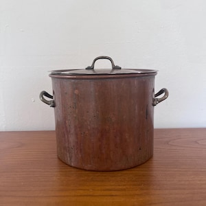 May include: A copper pot with a lid, featuring two side handles and a top handle. The pot has a cylindrical shape and a tarnished, aged appearance. The pot is sitting on a wooden surface.