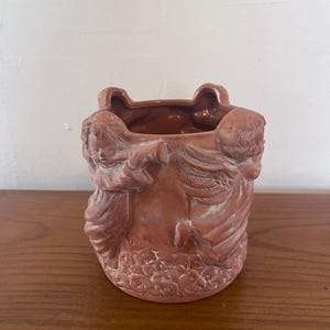 Puede incluir: Una maceta de cerámica color terracota con un relieve detallado de figuras de querubines. La maceta tiene forma cilíndrica con un borde festoneado y un motivo floral en la base.
