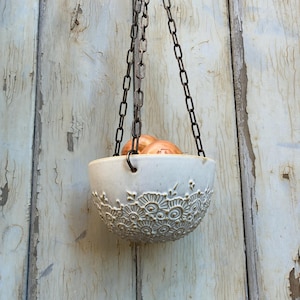 Könnte beinhalten: Ein hängender Keramik-Pflanztopf mit einem detaillierten floralen Muster am Rand. Die cremefarbene Schale wird von einer dunklen Metallkette gehalten und ist mit braunen Zwiebeln gefüllt. Der Hintergrund ist eine verwitterte, hellfarbige Holzwand.