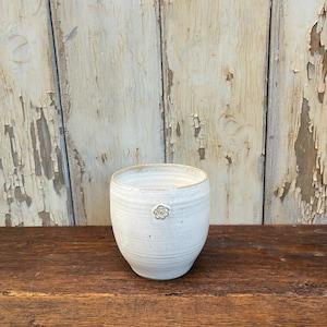 Könnte beinhalten: Ein weißer Keramikbecher mit einem kleinen Blumendesign. Der handgefertigte Becher hat eine abgerundete Form und eine strukturierte Oberfläche. Der Hintergrund ist eine Holzoberfläche und eine verwitterte weiße Wand.