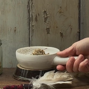 Könnte beinhalten: Eine weiße Keramikschale mit Griff, gefüllt mit getrockneten Kräutern. Die Schale steht auf einem Holzsockel, mit einem Glas Kräutern und einer Feder im Hintergrund. Die Schale hat ein florales Muster.