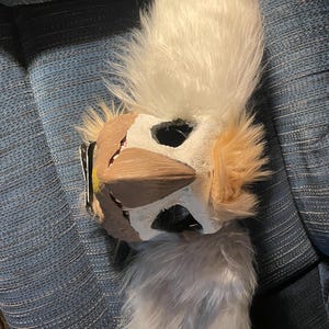 Può includere: Una maschera fatta a mano e pelosa con un design a forma di uccello. La maschera presenta un viso bianco con accenti marroni e beige, grandi fori per gli occhi e una cresta soffice di colore chiaro. La maschera è progettata per essere indossata.