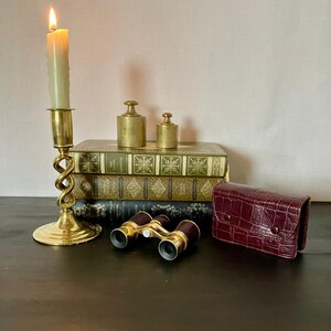 May include: A brass candlestick with a lit candle, two brass weights, three stacked hardcover books, a pair of brass and dark red opera glasses, and a dark red crocodile-embossed leather case arranged together on a dark surface.