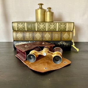 May include: A pair of vintage brass opera glasses with dark brown textured barrels and a central mother-of-pearl adjustment knob, resting in an open brown crocodile-patterned leather case in front of three stacked antique books and two brass weights.