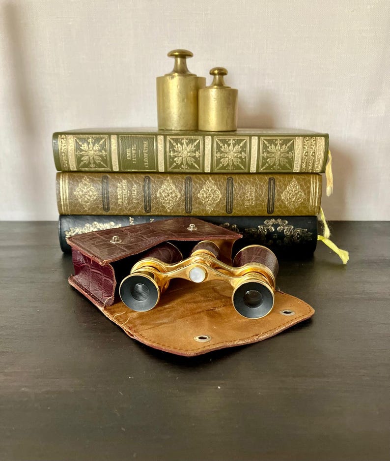 May include: A pair of vintage brass opera glasses with dark brown textured barrels and a central mother-of-pearl adjustment knob, resting in an open brown crocodile-patterned leather case in front of three stacked antique books and two brass weights.