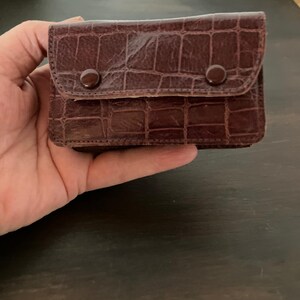 May include: A small, rectangular brown wallet featuring a textured crocodile-embossed pattern. The wallet has a front flap secured by two matching brown circular snap buttons, providing a structured and compact design for carrying small items.