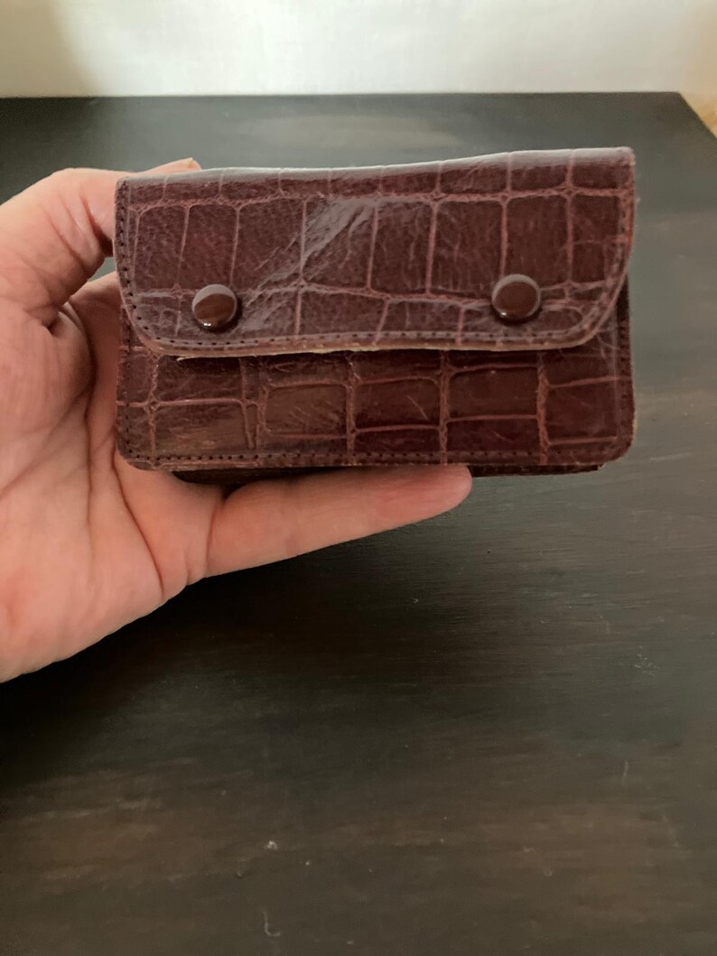 May include: A small, rectangular brown wallet featuring a textured crocodile-embossed pattern. The wallet has a front flap secured by two matching brown circular snap buttons, providing a structured and compact design for carrying small items.