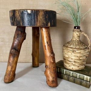 Op de afbeelding: Een rustieke houten kruk met drie natuurlijke poten. De ronde zitting is gemaakt van een plak van een boomstam, met donkere kleuring langs de randen. Een geweven fles en twee groene boeken maken de natuurlijke, aardse esthetiek compleet.