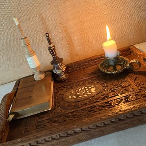 Può includere: Un vassoio rettangolare in legno con intagli floreali complessi e un motivo ovale centrale intarsiato. Sul vassoio si trovano un piccolo libro marrone, due sottili fusi decorativi in legno e un portacandele in ottone con una candela bianca accesa.