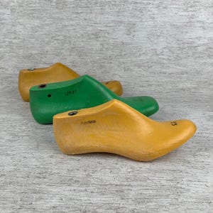Può includere: Tre forme per scarpe in legno nei toni del giallo e del verde sono disposte su una superficie grigio chiaro e bianca. Le forme delle scarpe hanno una forma liscia e curva, con segni visibili, tra cui i numeri "12027" e "35".