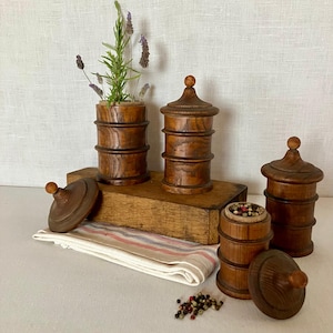 Puede incluir: Un conjunto de cuatro recipientes cilíndricos de madera con tapas, con un acabado de veta de madera marrón oscuro. Dos recipientes están cerrados, uno contiene ramas verdes y otro está abierto, revelando granos de pimienta coloridos en su interior.