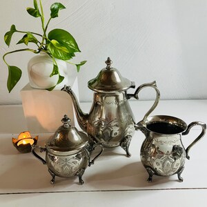 Può includere: Servizio da tè in argento antico con disegni floreali ornati. Il set include una teiera, una zuccheriera e una lattiera, ciascuna con manici e piedini decorativi. Un piccolo vaso bianco con foglie verdi è sullo sfondo.