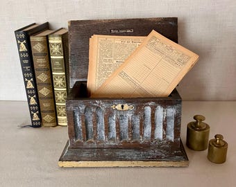 May include: A dark brown wooden box with a distressed white finish and a central keyhole, containing several aged, tan-colored paper documents. To the left are three vintage books with gold-embossed spines, and to the right are two brass weights.