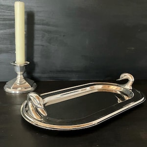 Op de afbeelding: Een zilverkleurig ovaal dienblad met twee zwanenvormige handvatten aan de uiteinden. Naast het dienblad staat een zilverkleurige metalen kandelaar met een zeshoekige voet, waarin een enkele, hoge, gebroken witte cilindrische kaars staat.
