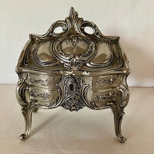 May include: A silver-toned metal jewellery box shaped like a miniature chest of drawers. The piece features an ornate, raised relief design with curved legs, a central keyhole, and decorative drawer pulls on the front, topped with a curved lid.