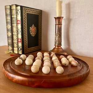 May include: A round wooden board game featuring a set of smooth, cream-colored stone spheres arranged in a pattern. The game is accompanied by three leather-bound books with gold detailing and a tall, metallic candle holder with a cream candle.