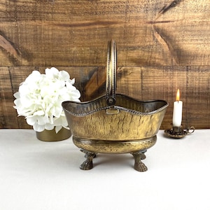 Peut inclure: Un panier en laiton vintage avec une poignée, une surface texturée et des pieds en forme de griffes. Le panier est présenté avec un arrangement floral blanc et une bougie allumée dans un petit support, sur fond de bois.
