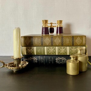 May include: A stack of three hardcover books with gold and black spines, topped with a pair of burgundy and gold opera glasses. Beside the books sits a brass candle holder with a cream-colored candle and two cylindrical brass weights.