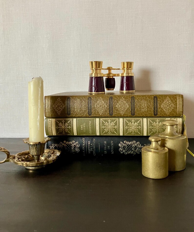 May include: A stack of three hardcover books with gold and black spines, topped with a pair of burgundy and gold opera glasses. Beside the books sits a brass candle holder with a cream-colored candle and two cylindrical brass weights.
