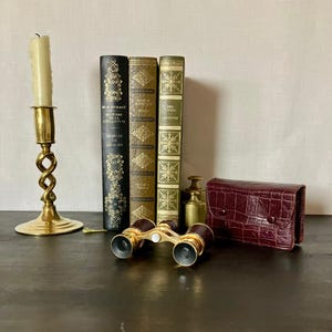 May include: A brass candlestick with a white candle, three hardcover books with gold-embossed spines, a pair of brass opera glasses with a central white focus knob, two small brass weights, and a dark red crocodile-textured leather case.
