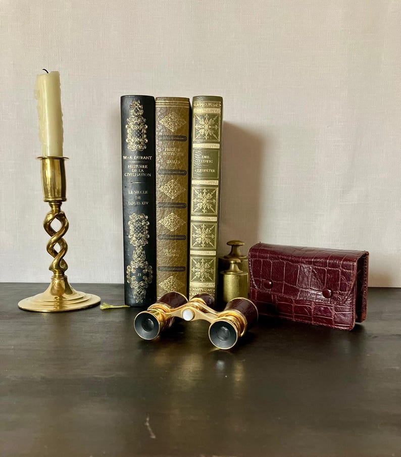May include: A brass candlestick with a white candle, three hardcover books with gold-embossed spines, a pair of brass opera glasses with a central white focus knob, two small brass weights, and a dark red crocodile-textured leather case.