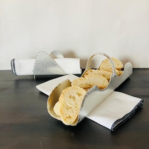 May include: A silver-toned metal bread basket with a curved handle containing sliced bread, accompanied by a matching silver-toned metal napkin holder. Both items are paired with white cloth napkins featuring a dark grey stitched edge detail.