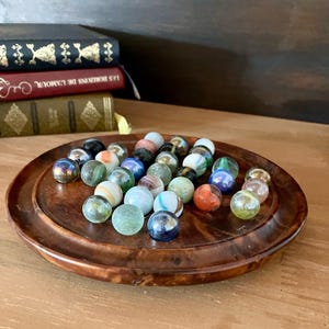 Puede incluir: Una colección de pequeñas canicas de vidrio esféricas en varios colores, incluyendo azul, verde, blanco y naranja, sobre una bandeja circular de madera pulida con un borde elevado. Las canicas presentan diversos patrones, remolinos y acabados translúcidos.