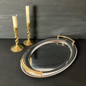 Könnte beinhalten: Ein ovales silberfarbenes Serviertablett mit goldfarbenen Griffen, begleitet von zwei Messingkerzenhaltern mit gedrehten Stielen, die jeweils eine cremeweiße Stabkerze halten. Das Tablett hat eine reflektierende Oberfläche und geschwungene goldfarbene Griffe.