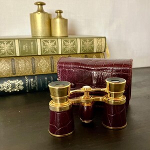 May include: A pair of opera glasses featuring a dark burgundy, crocodile-textured leather finish on the barrels and a matching hard-shell carrying case. The optical instrument is accented with polished gold-tone metal hardware and a central focus knob.