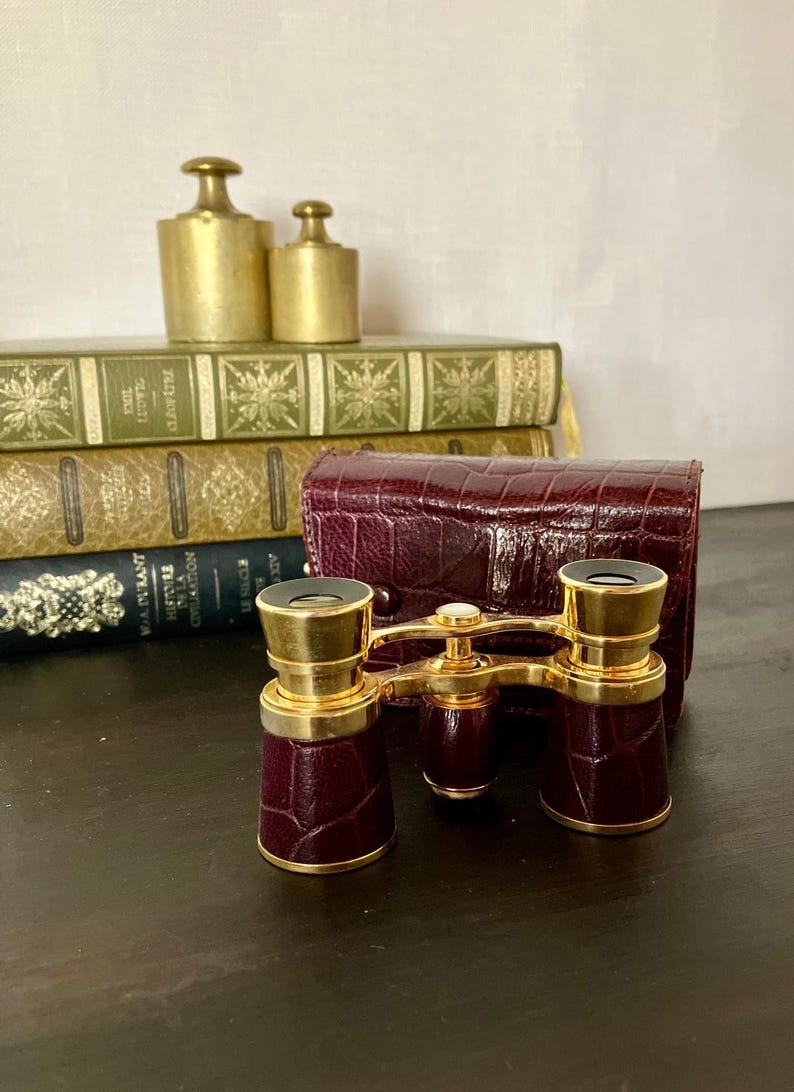 May include: A pair of opera glasses featuring a dark burgundy, crocodile-textured leather finish on the barrels and a matching hard-shell carrying case. The optical instrument is accented with polished gold-tone metal hardware and a central focus knob.