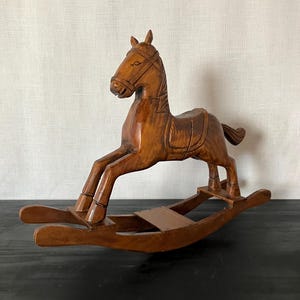 Könnte beinhalten: Ein braunes Schaukelpferd aus Holz mit geschnitzten Details wie Zaumzeug, Sattel und einer fließenden Mähne. Das Pferd ist auf zwei geschwungenen Kufen montiert, die durch ein zentrales Holzbrett verbunden sind, mit polierter Oberfläche und Maserung.