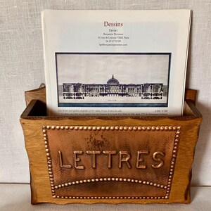 以下が含まれることがあります： 「LETTRES」という文字と装飾的な鋲が施された中央の銅製パネルを持つ木製の壁掛けレターホルダー。建物が描かれた「Dessins」というタイトルの白い書類が中に差し込まれています。