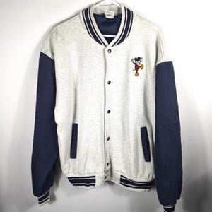 May include: A gray and navy blue varsity jacket with a button-up front. The jacket features navy blue sleeves, pockets, and trim, with a white collar and cuffs. A Mickey Mouse graphic is embroidered on the chest.
