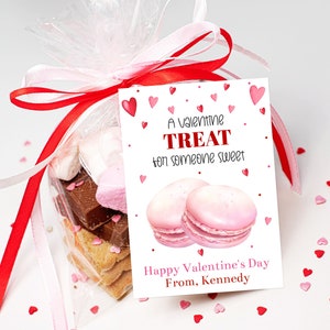 May include: A Valentine's Day treat bag with a pink and red ribbon, filled with cookies, chocolate, and marshmallows. The bag has a tag that says "A Valentine Treat for someone sweet. Happy Valentine's Day From, Kennedy."