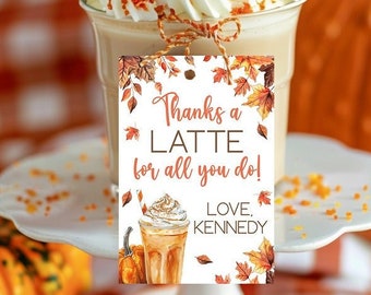 May include: A latte with whipped cream and orange sprinkles sits on a white cake stand. A gift tag with fall leaves and the words "Thanks a Latte for all you do!" is attached. Pumpkins and a checkered tablecloth complete the autumn scene.