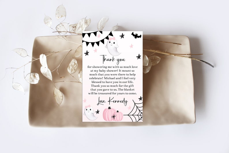 May include: A baby shower thank you card with a ghost, pumpkins, and spiderweb design. The card includes the text "Thank you" and a personalized message. The card is on a beige tray with decorative sprigs.
