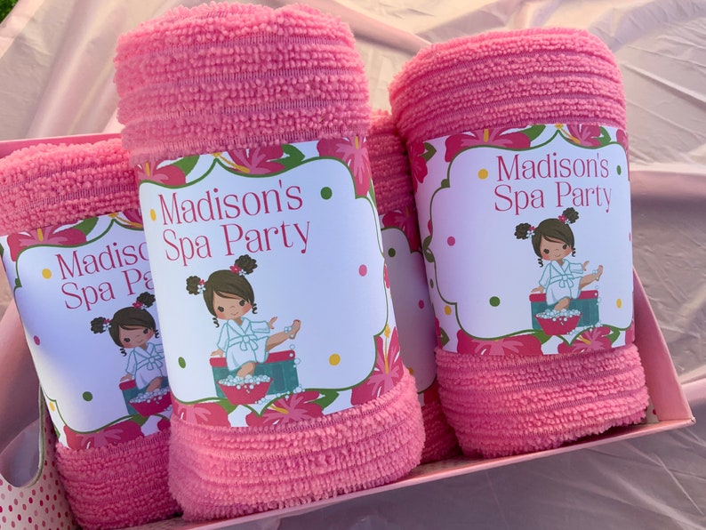 K&ouml;nnte beinhalten: Zwei rosa Handt&uuml;cher mit floralen Etiketten, auf denen "Madison's Spa Party" steht und ein Cartoon-M&auml;dchen in einem Bademantel abgebildet ist.