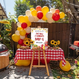 May include: A welcome sign for a baby shower with a barbecue theme. The sign is white with red and yellow accents and says "Welcome to Matthew & Kennedy's Baby-Q May 30, 2024". The sign is decorated with sunflowers, hot dogs, hamburgers, and other barbecue-themed items.