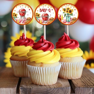 May include: Three yellow and red cupcakes with white liners topped with a cupcake topper. The toppers are round and feature a watercolour illustration of a grill, sunflowers, and the text "BABY-Q".
