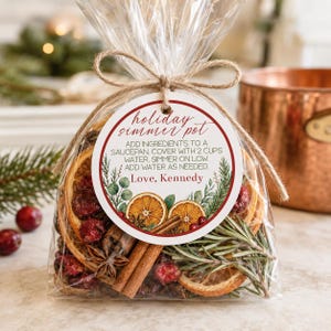 May include: A clear plastic bag filled with dried orange slices, cinnamon sticks, star anise, cranberries, and rosemary sprigs. A round white tag with a red border is tied with twine, reading: holiday simmer pot, instructions, and Love, Kennedy.