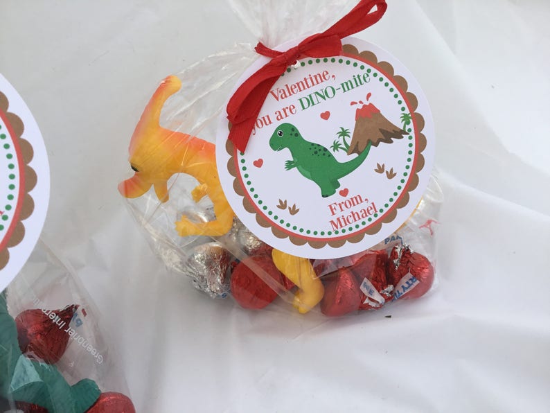 Puede incluir: Una bolsa de golosinas para el D&iacute;a de San Valent&iacute;n con una etiqueta roja, blanca y marr&oacute;n con tem&aacute;tica de dinosaurios que dice "Valentine, you are DINO-mite. From, Michael". La bolsa contiene caramelos de chocolate rojos y amarillos y un peque&ntilde;o juguete de dinosaurio amarillo.