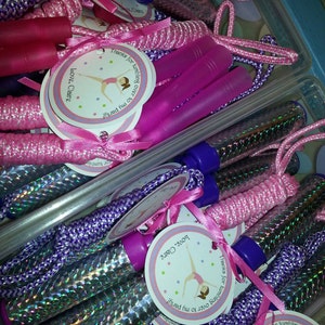 K&ouml;nnte beinhalten: Rosa und lila Springseile mit silbernem Glitzer-&Uuml;berzug und wei&szlig;en Etiketten mit der Aufschrift "Danke, dass du zu meiner Party gekommen bist! Liebe, Claire".
